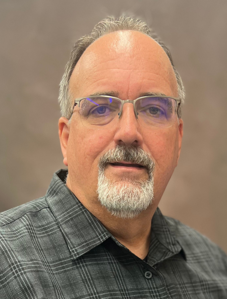 A man with short gray hair and a beard, wearing glasses and a gray plaid shirt, poses against a neutral background.